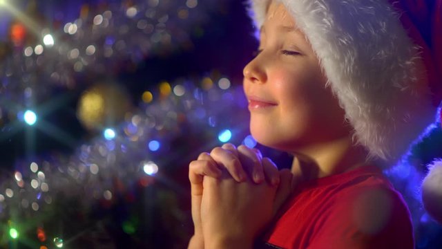 4K video of a smiling boy  is praying near Christmas tree about christmas gifts. Boy in a Santa hat is dreaming Christmas gifts on  Christmas.  Happy 8 year old child is preparing for the New Year.
