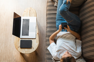 Woman use a smartphone while lying on a sofa with a laptop and ereader on a table. Studying online,...