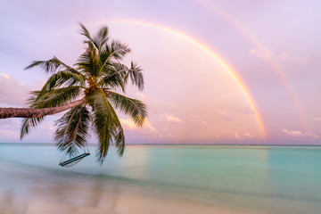 Landscape of paradise tropical island beach, sunrise sunset with beautiful rainbows over calm sea water. Wonderful scenery, tropical beach evening view. Palm tree with beach swing or hammock