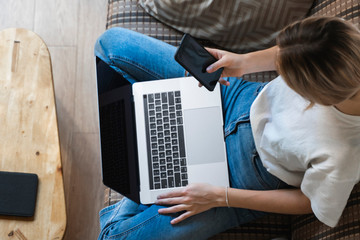 Woman with a laptop is sitting on a sofa and use a smartphone. Study and work online, freelance....