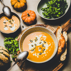 Autumn, winter warming seasonal meal. Flat-lay of pumpkin soup with seeds, parsley and cream in bowls over grey concrete background, top view, square crop. Vegetarian, healthy, comfort food concept