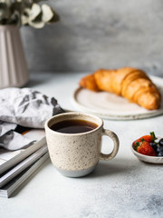 Delicious breakfast - the cup of coffee, croissant, berries and various magazins on the table at the grey background. Copy space.