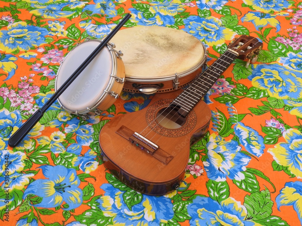 Wall mural Three Brazilian musical instruments: cavaquinho, pandeiro (tambourine) and tamborim with drumstick on a colorful “chitao” fabric with floral prints. They are widely used in samba and pagode ensembles.