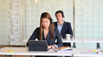 Beautiful call center woman in black suit she looking on tablet screen and sitting at work desk in front of her colleague in the modern office.