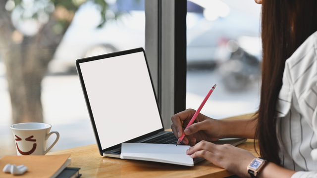 Close-up image of young beautiful confident girl taking notes in front of white blank screen laptop on the wooden cafe bar. Modern cafe concept.