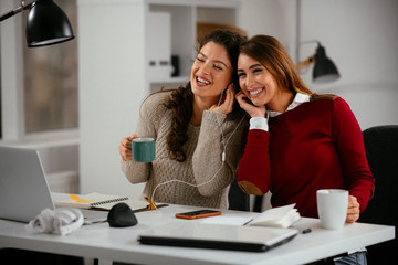 Two beautiful businesswomen having fun in office. Colleagues listening music together.