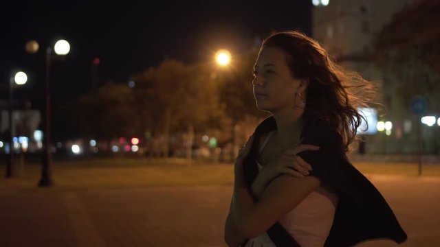 Beautiful girl at night in the city, the wind blows her hair. Elegance of a girl, arms folded on her chest. Girl in a dress and jacket on her shoulders.
