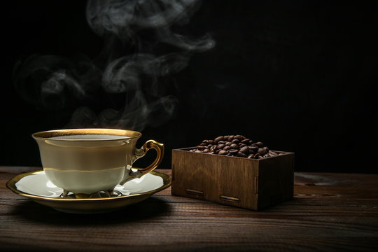 Cup Of Hot Coffee On A Wooden Surface On A Black Background. Background With Roasted Coffee Grains. Steam From A Cup Of Coffee