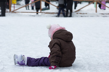 Fototapeta na wymiar Side view of caucasian child of three years old sitting on the rink with skates on legs in wintertime