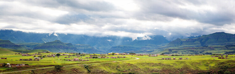 Fototapeta na wymiar Panorama of a village in the mountains of Maloti Drakensberg Park with sunbeams breaking through the clouds and illuminating the green meadows, South Africa