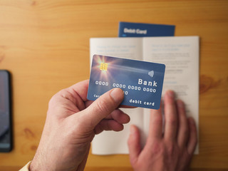 Man holding debit bank card in left hand in focus, terms and conditions booklet on a a wooden table out of focus, right hand over the page. Concept managing financial information and technology.
