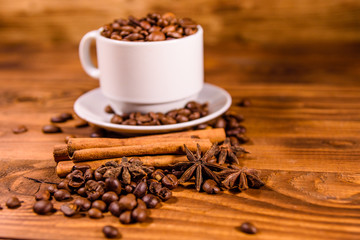White cup filled with coffee beans, star anise and cinnamon sticks on wooden table