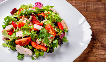 Fresh salad with chicken breast, arugula, black olives,red pepper, lettuce, fresh sald leaves and tomato on a white plate on wooden table background