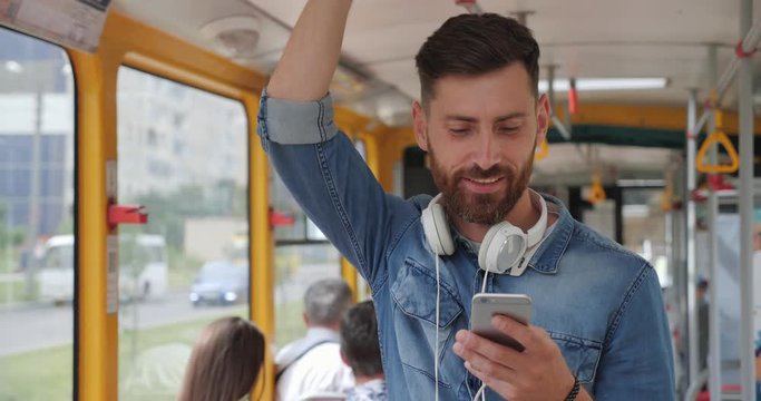 Casually dressed Attractive man Using tram, Holding Smartphone with one hand, handrail with other. Looking smiling and gorgeous. Having good mood and positive emotions on his face