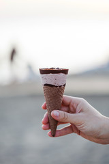 Cream ice cream with chocolate in a waffle cone close-up in your hand on the background of the sea and sandy beach.