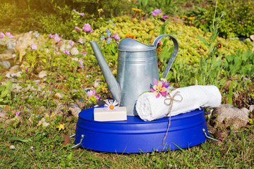 Metal watering can, purple basin, toilet soap and white towel with flowers in garden. Bath tools. Summer out of town