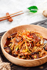Pan-Asian food. Noodles with vegetables, shiitake mushrooms and meat in a beautiful wooden plate on a dark rustic tray on a light concrete background. Beautiful composition. Close-up. Space