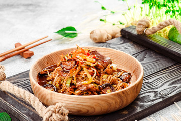 Pan-Asian food. Noodles with vegetables, shiitake mushrooms and meat in a beautiful wooden plate on a dark rustic tray on a light concrete background. Beautiful composition. Close-up. Space