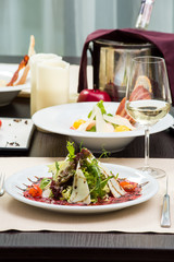 Fresh salad with beef carpaccio, parmesan, tomatoes and sauce on a white plate and a glass of white wine on a table in a restaurant. Close-up. Space