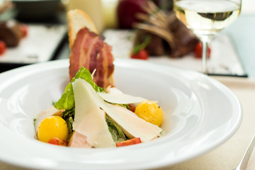 Traditional Caesar salad with chicken breast, tomatoes, lettuce, egg, parmesan and croutons in a white plate and a glass of white wine on a table in a restaurant. Close-up