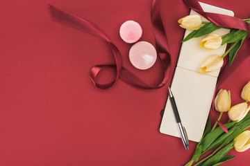 top view of tulips with silk ribbon, macarons and empty notebook with pen isolated on red