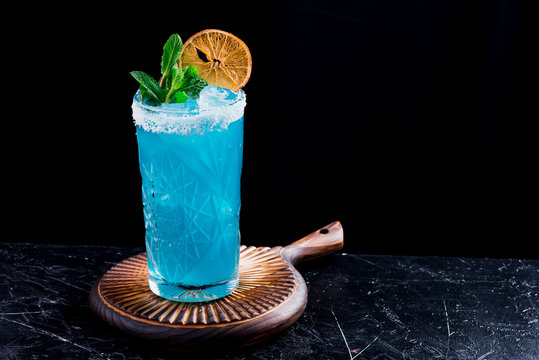Blue cocktail with ice, decorated with mint and slice of dry orange in beautiful faceted glassm on black background