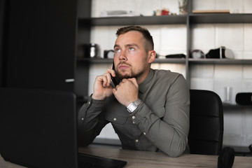 Young businessman or student in a shirt sitting against monitor of computer and speaking with his business partner by smartphone. Working on a pc at a table in the office with a thoughtful expression.