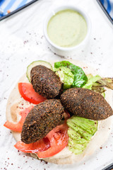 National Israeli dish - Pita with falafel, lettuce, tomato slices and sauce on a white square plate, on a dark wooden background decorated with legumes and with a blue napkin. Close up. Space