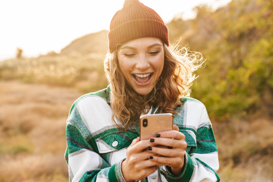 Image of joyful young woman holding cellphone while walking outdoors