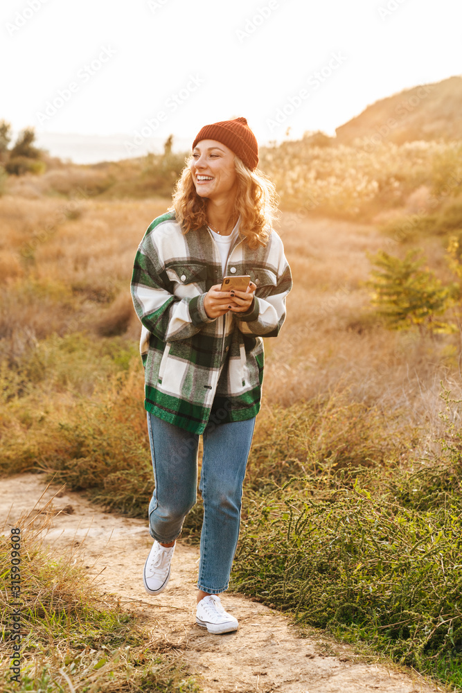 Wall mural image of joyful young woman holding cellphone while walking outdoors