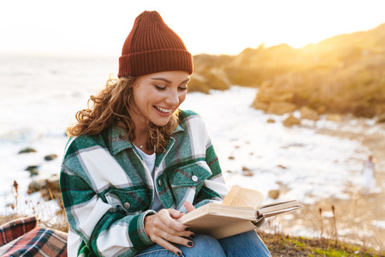 Image of joyful young caucasian woman reading book by seaside