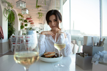 Adult beautiful woman sit in elite restaurant with a beautiful view of the sea, her order seafood and drink. The female person wearing white shirt. Girl chosses the italian wine and mussels.