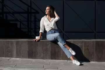 A brunette woman with long hair in fashionable in blue jeans and a white shirt posing outdoors.Fashion photo concept. Woman near business center.Beautiful female business center worker near her office