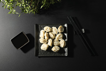 Square plate with dumplings, chopsticks, plant and soy sauce on black background, top view