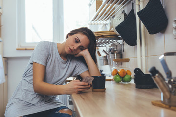 heartbroken woman using mobile phone, sitting in her kitchen drinking coffee