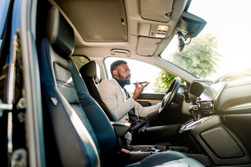 Young successful African American businessman talking on speakerphone through microphone with client, sitting in the expensive car. Negotiations and business meetings.