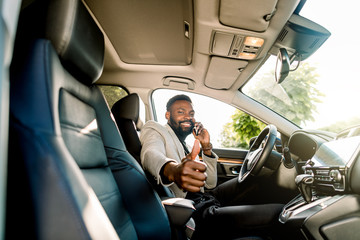 Successful handsome African man businessman and boss sitting in the car, talking on the phone, smiling, looking at camera and showing thumb up. Business concept
