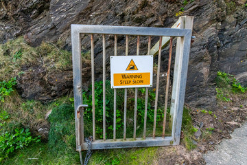 A steep slope warning sign on a metal gate on a cliffside 