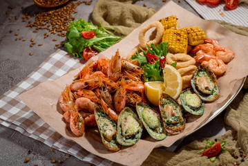 Beer snack: fried onion rings, fried mussels in shells, fried corn and prawns on parchment on concrete background, decorated with checkered napkin with fresh vegetables and beautiful bread. Space