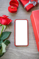 White blank screen of phone with gift boxes, rose and ring on wooden background.