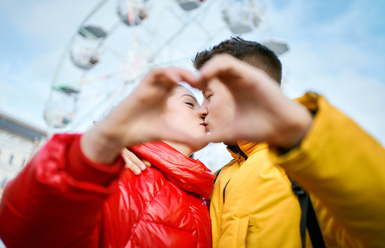young romantic couple on date, embracing and kissing through a heart gesture made with their fingers, spending time together in the city. Lovers wearning in bright yellow and red down jackets. ferris