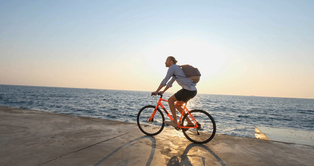 Young handsome male in casual wear ride on the colorful bicycle on the morning beach against beautiful sunset and the sea