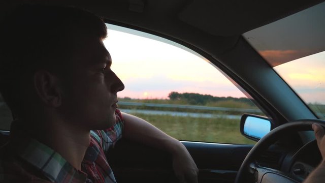Profile of handsome man in shirt driving car through countryside. Young guy riding on auto with his hand out of window and enjoying road trip. Beautiful evening view at background. Slow motion Closeup