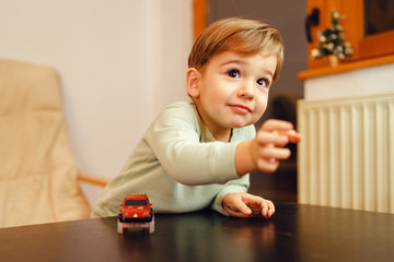 Portrait of small little caucasian boy two years old playing with plastic car toys at home by the table reaching toward leaning