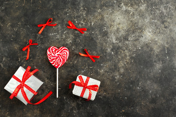 White-red striped lollipop in the form of a heart and festive wrapped gifts on a dark aged background, flat lay. Around the candy are red bows. Valentines day concept. Love for shopping, copy space