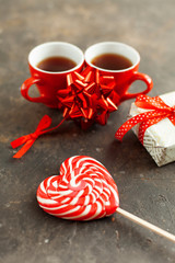 Two red cups of hot espresso or coffee, beautiful wrapped gift and decorative bow on a dark textured background. White-red lollipop in the form of a heart closeup. Valentines day romantic date concept