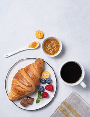 Fresh  croissant on a white plate with kumquat jam, blueberries and raspberries with a Cup of  vegan coffee on a gray background.