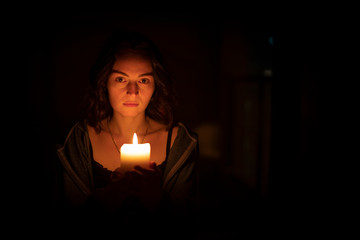 The mysterious religious orthodox brunette girl with brown eyes looks piercingly, with a serious look, holding a bright, lighted candle in folded hands. Horizontal view