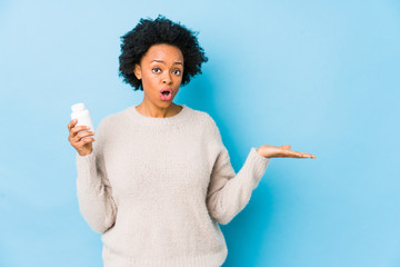 Middle age african american woman holding a vitamin bottle impressed holding copy space on palm.