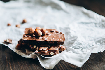 Milk chocolate with nuts: peanuts, hazelnuts on white parchment paper with a nutcracker and on a dark wooden table. Selective focus